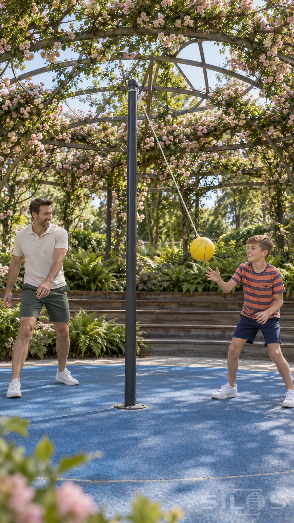A father and son smile during tetherball on a blue court beneath a rose pergola, with one yellow ball hanging from a taut cord.