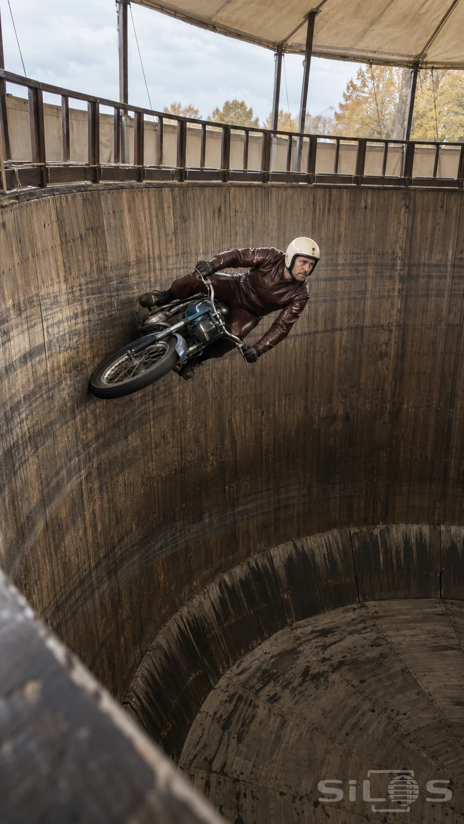 An adult motorcycle performer in burgundy leathers rides high on the vertical wall of a large wooden fairground drum.