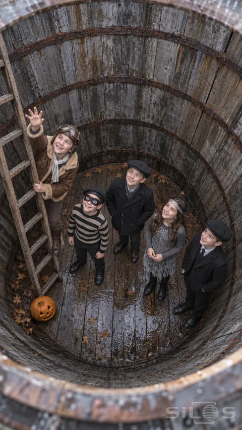 Five costumed children look upward inside an empty wooden tank; with an aviator boy holding a ladder beside four friends.