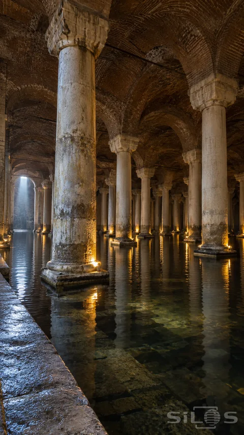 Vaulted cistern reflections spread across dark water beneath stone columns and brick arches with warm lamps at their bases.