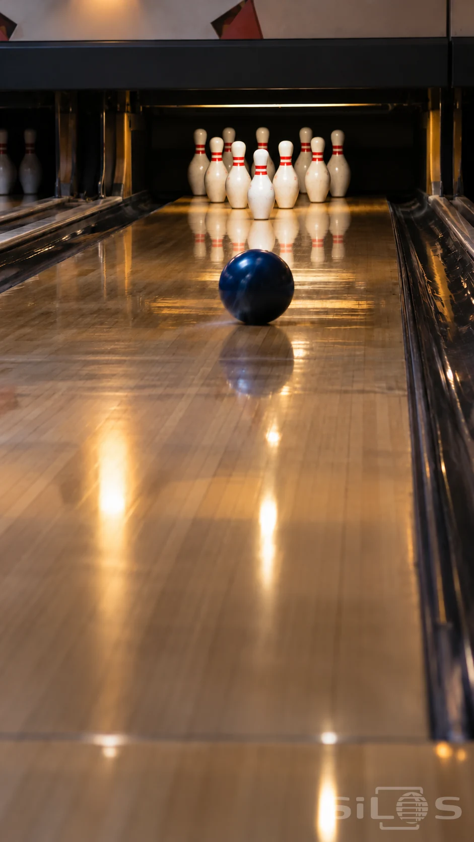 A blue bowling ball rolls along a polished lane toward ten white pins with red neck bands arranged in a triangular rack.