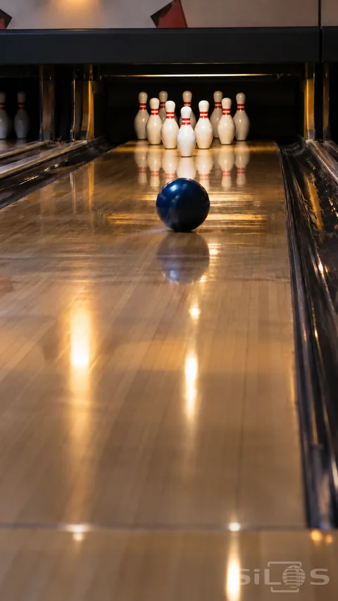 A blue bowling ball rolls along a polished lane toward ten white pins with red neck bands arranged in a triangular rack.