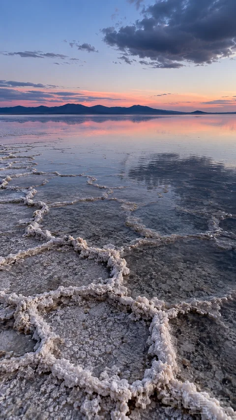 Raised white salt polygons fade into shallow rainwater that reflects a rose horizon; distant hills; and a dark cloud in a blue evening sky.
