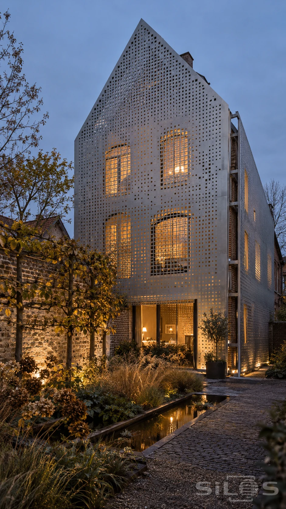 Tall gabled house wrapped in perforated silver metal panels with warm windows behind, beside a walled garden and narrow reflecting pool