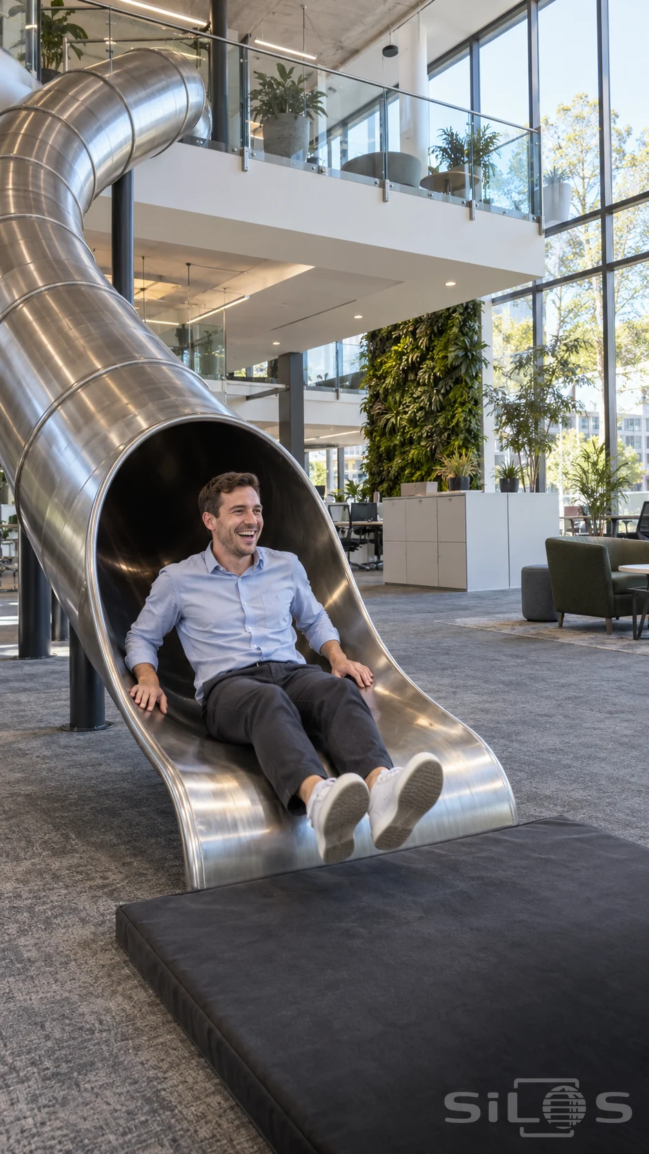 A man in a pale blue shirt laughs as he reaches the bottom of a steel office slide above a dark landing mat.