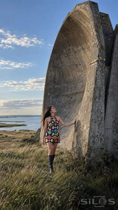 A woman in a colorful crochet dress and black boots walks beside a large concrete sound mirror in a coastal meadow.