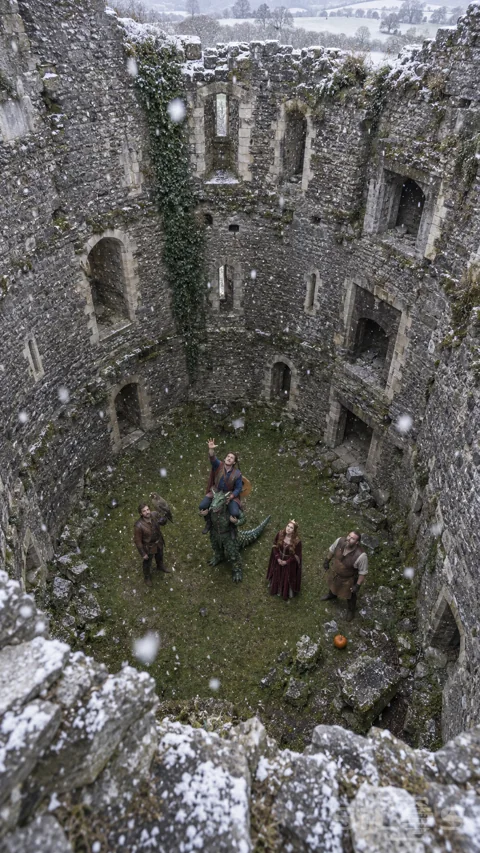 Five costumed adults look up from the grassy floor of a ruined keep as snow falls.