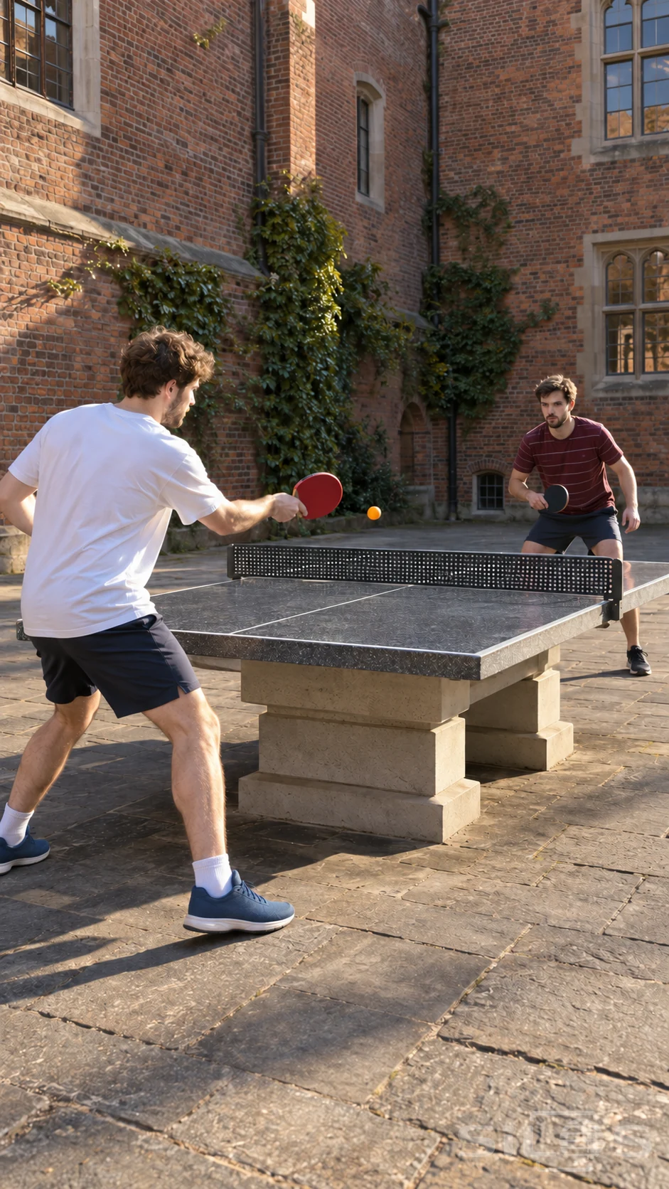 Two men trade a table-tennis rally across a concrete table with an orange ball above its fixed metal net in a sunny brick courtyard.