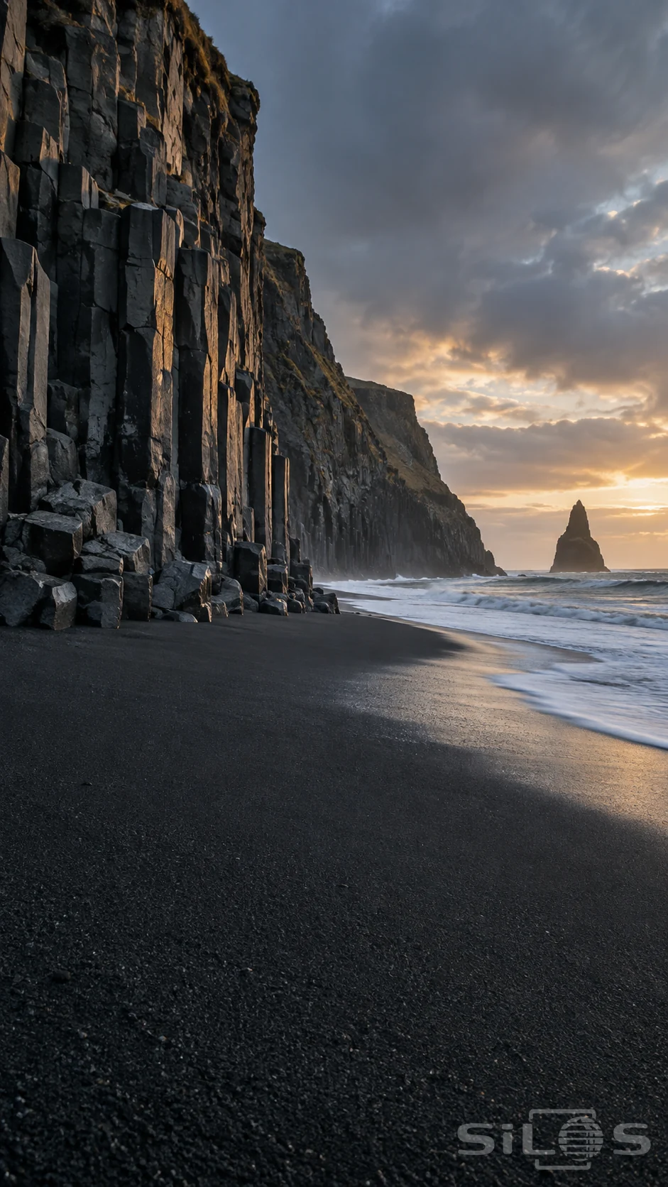 Tall basalt columns rise above wet black sand while white surf curves toward a distant sea stack in pale dawn light.