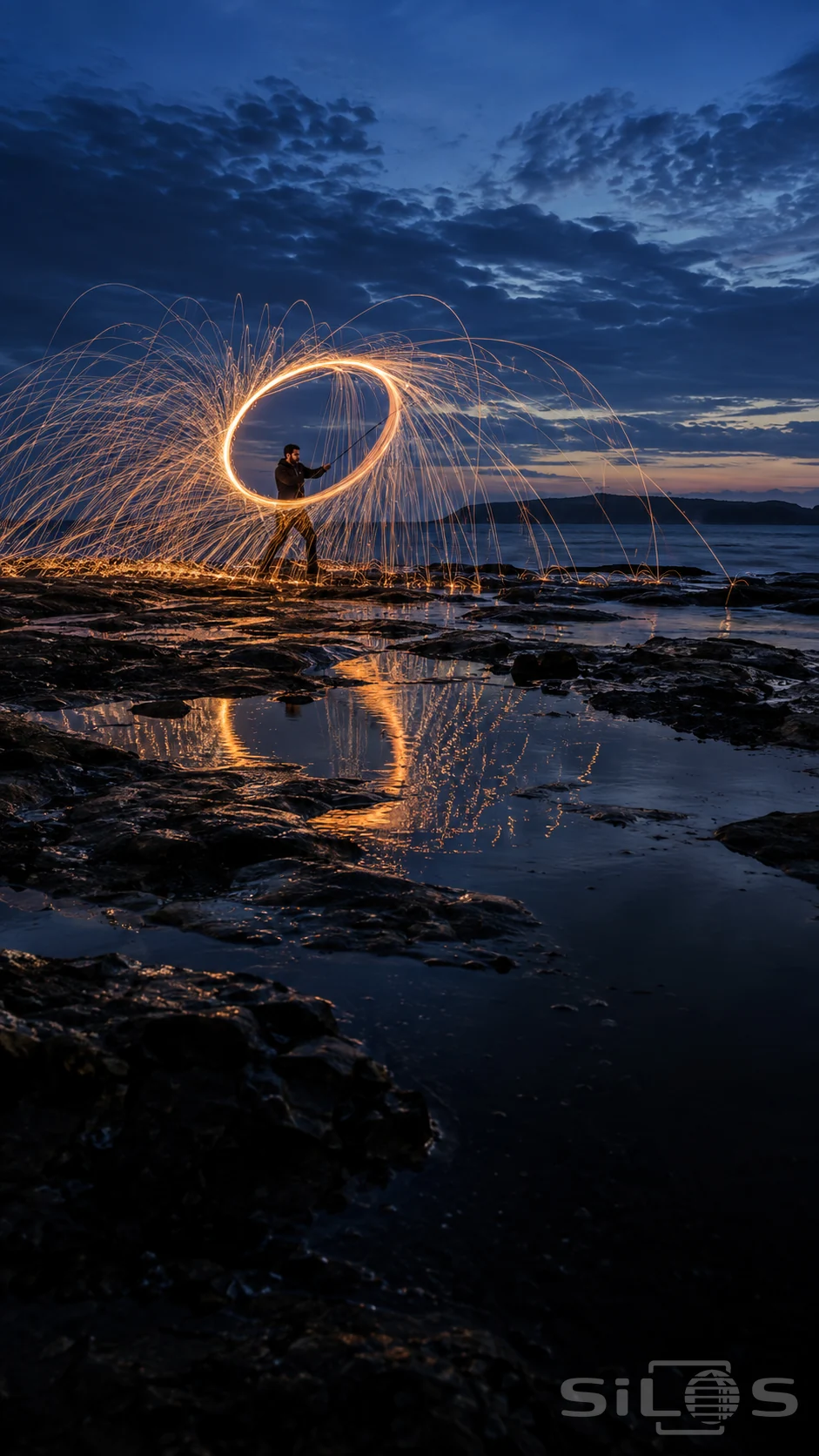 Spark light painting with a golden circular trail around an adult performer on wet rock, reflected beneath a blue-hour sky.