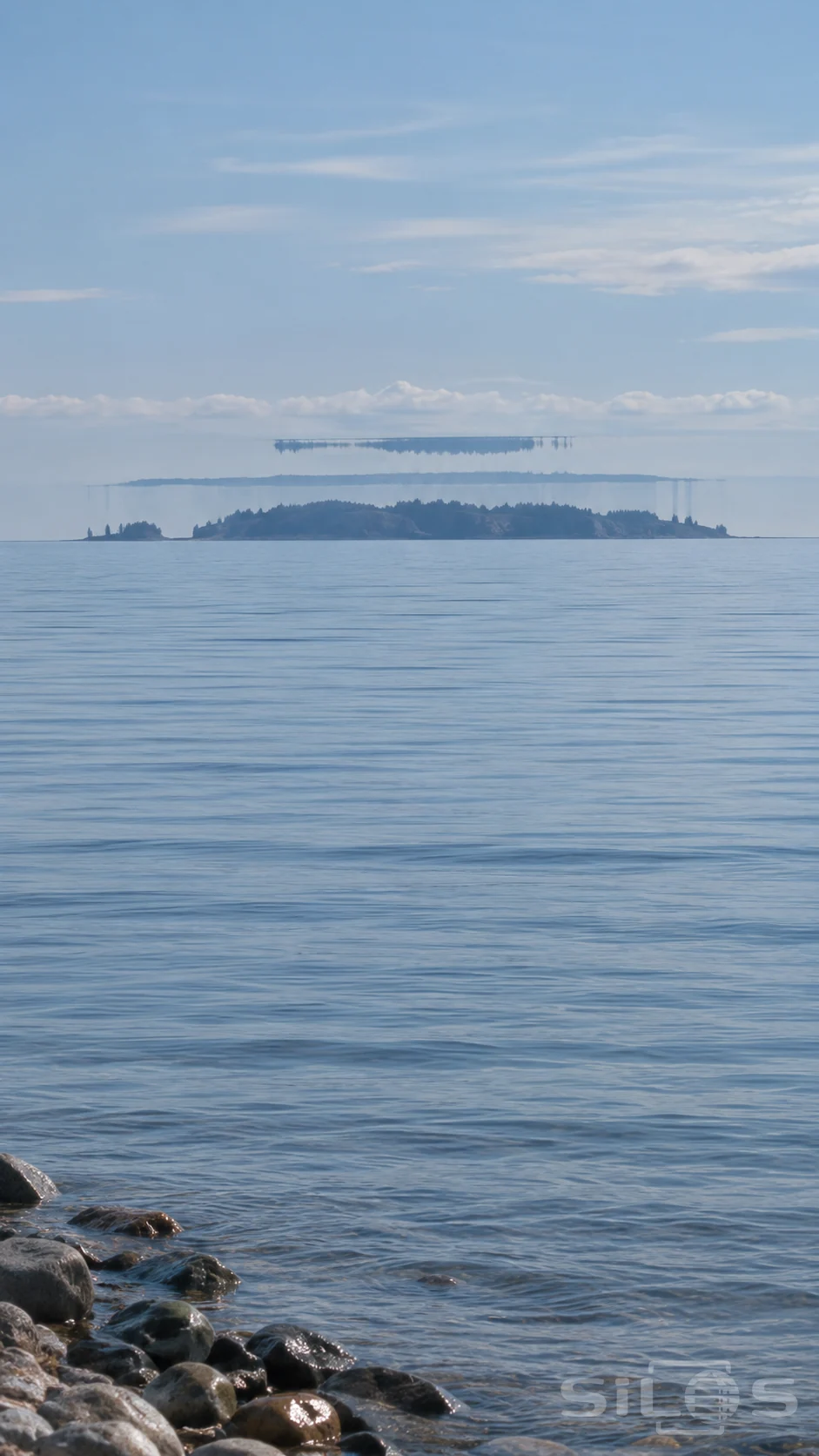 Mirage horizon with stacked island silhouettes above calm blue water and wet shoreline stones.