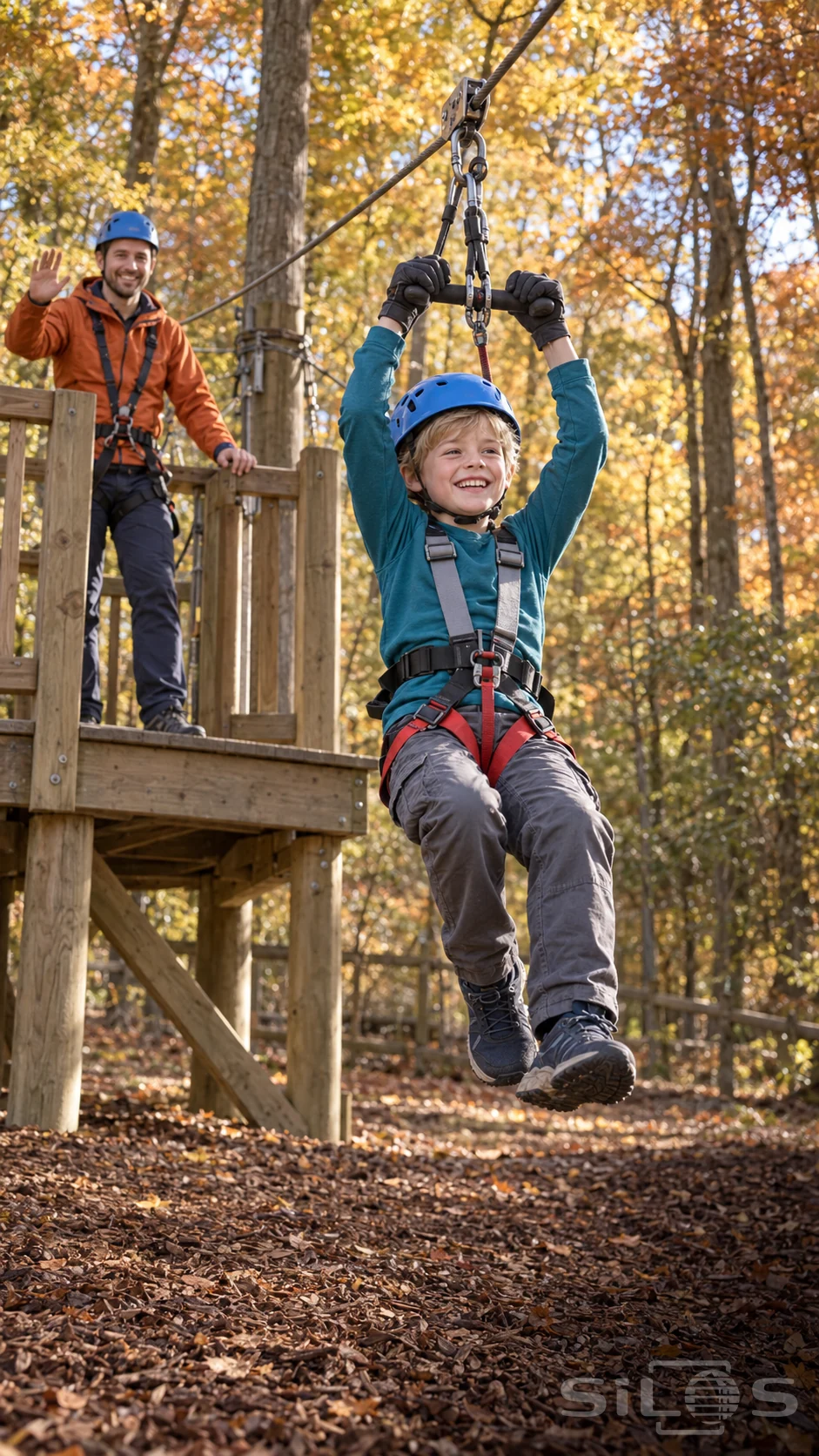 A boy in a blue helmet and full-body harness glides on a low zipline while his father waves from a timber platform.