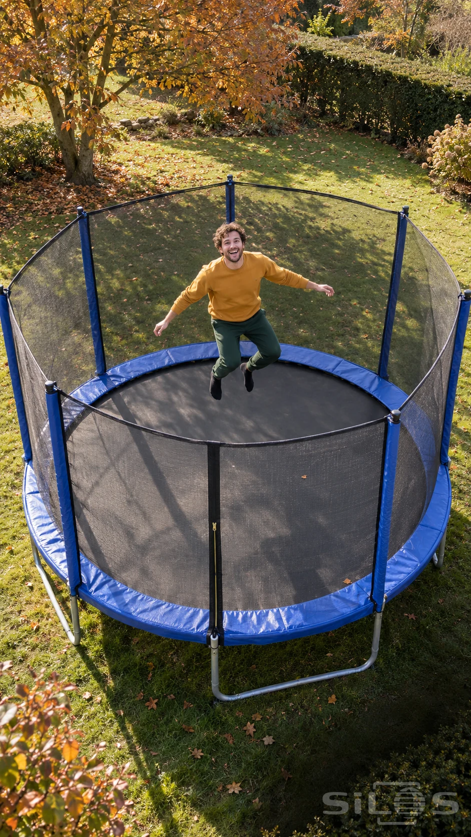 A man in a mustard sweatshirt jumps on a round trampoline enclosed by a black safety net in an autumn garden.