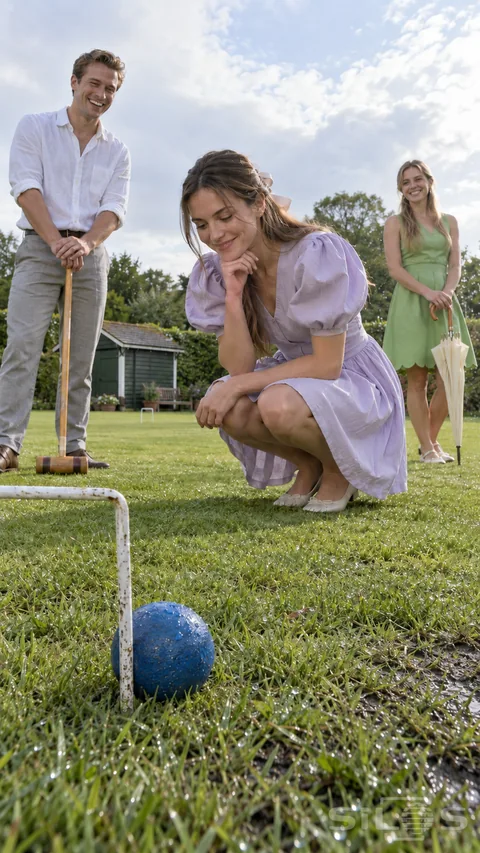 Croquet lawn moment with a woman crouching beside a blue ball at a white hoop while two friends watch with a mallet and closed umbrella.