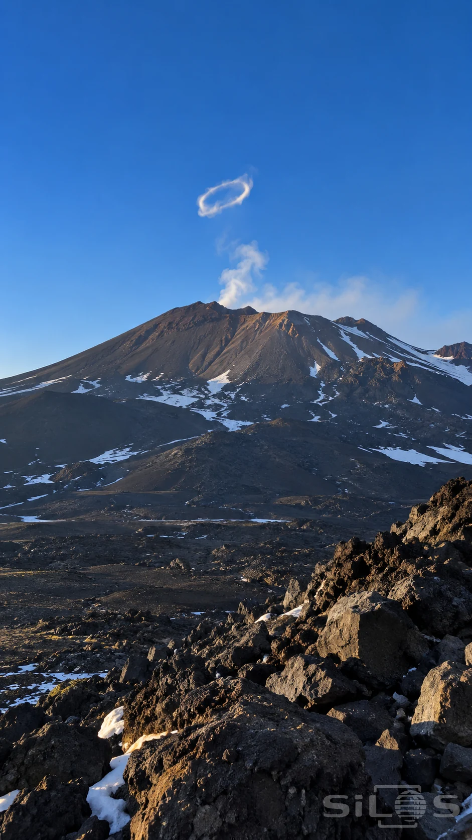 A volcanic vapor ring hangs above a dark summit and faint plume with patches of snow between rough lava stones.