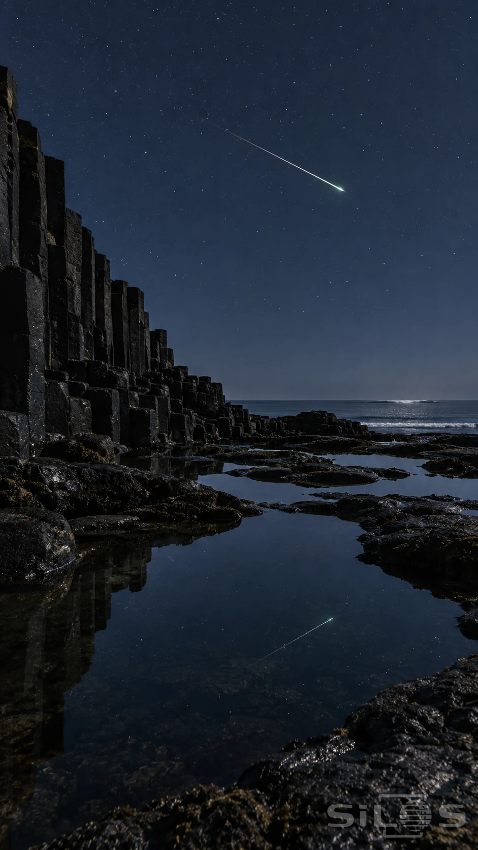 A thin meteor streak is reflected in quiet tidal pools beside tall dark basalt columns beneath a starry blue night sky.