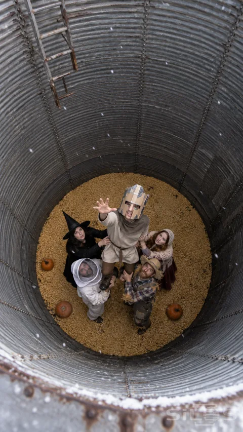 Overhead view of five costumed adults supporting a reaching knight inside a tall steel grain silo.
