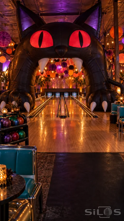A giant inflatable black cat arch stands above bowling lanes beneath colorful paper decorations and warm lights.