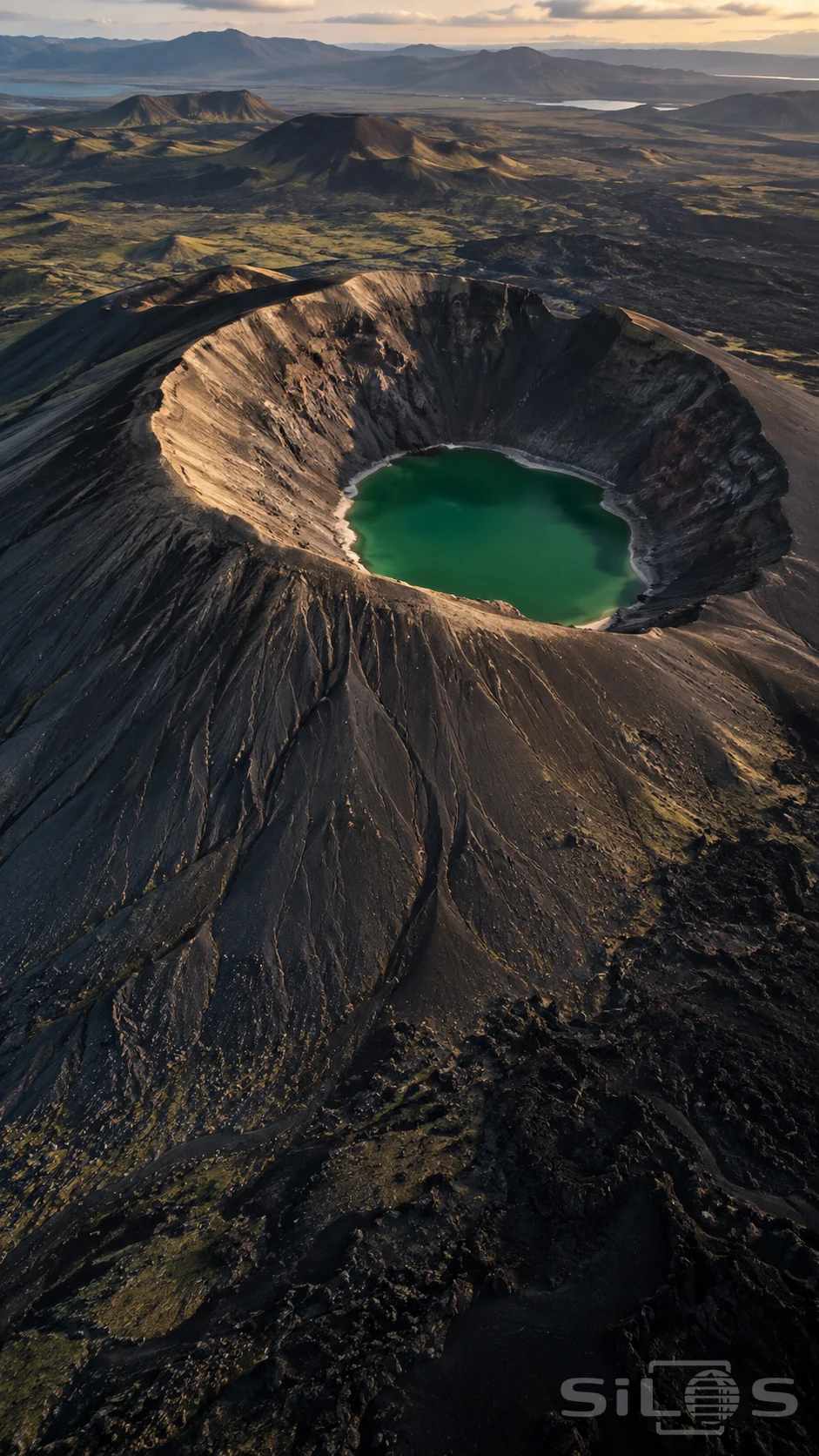 An emerald pool fills the bottom of a dark volcanic crater, with deeply grooved slopes and smaller cones across the distant plain.