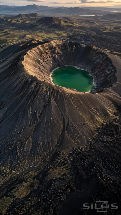 An emerald pool fills the bottom of a dark volcanic crater; with deeply grooved slopes and smaller cones across the distant plain.