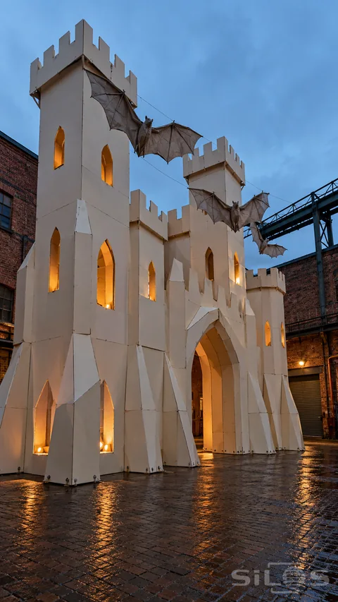 A tall Halloween paper castle with lit windows and hanging cloth bats stands in a wet brick loading yard at twilight.