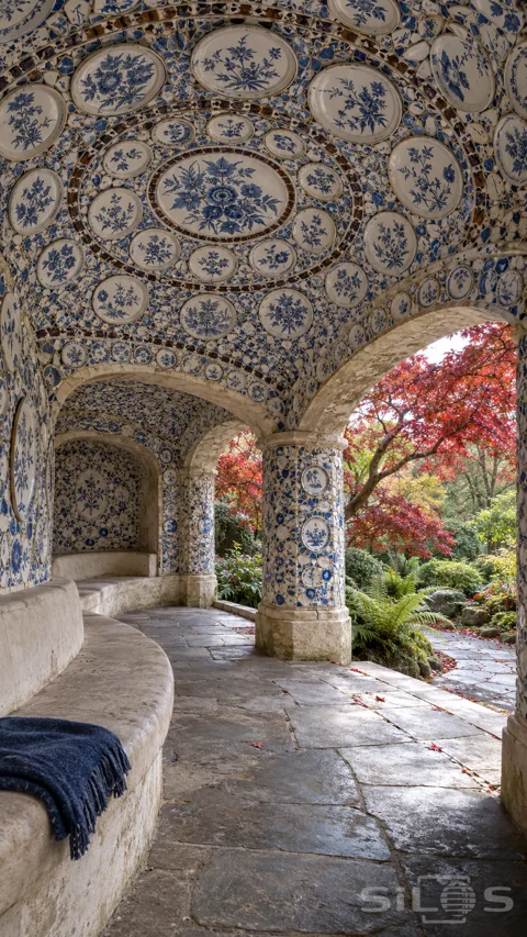 Blue and white plate mosaics cover an arched garden shelter with a curved stone bench; red maple leaves and a wet path.