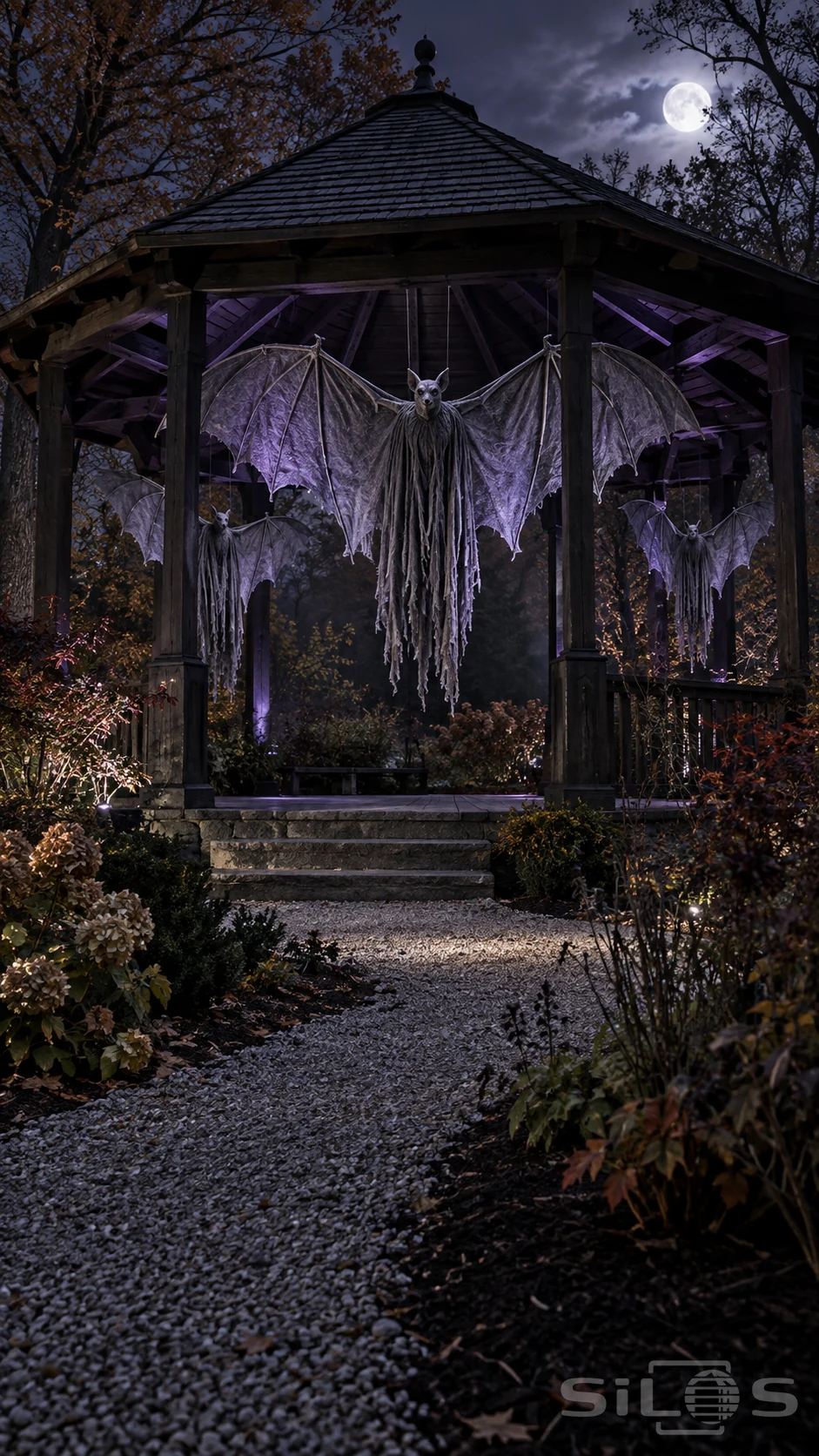 Three bat props with gray wings and ragged hanging cloth are suspended inside a timber gazebo lit violet beneath the moon.