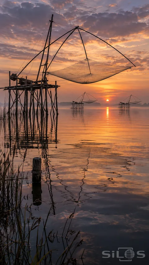 Lift nets hang from arcing bamboo arms above timber platforms; reflected in a calm lagoon beneath warm dawn clouds.