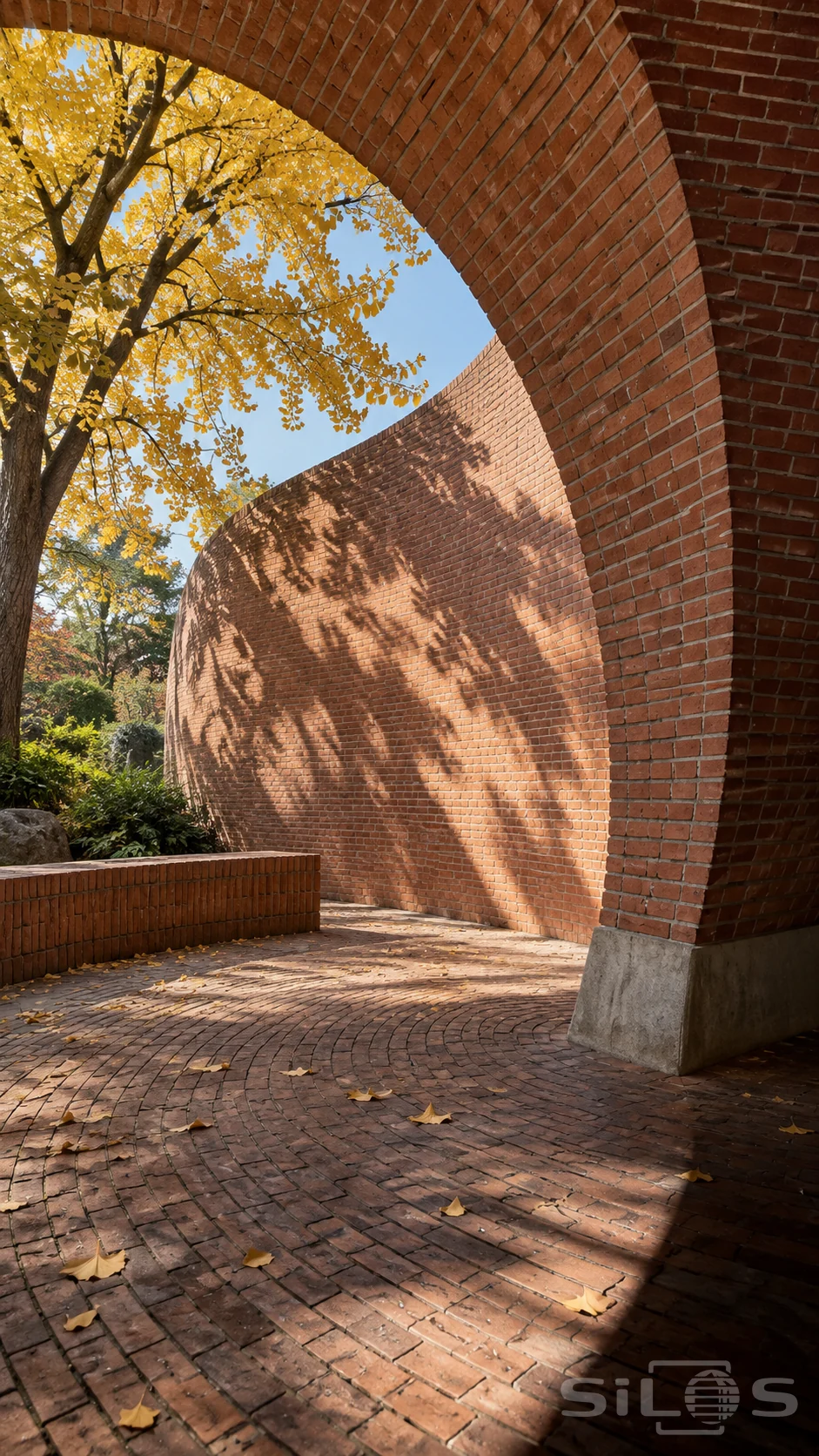 A curved brick shell and tall arch enclose an empty paved courtyard with a low bench and yellow leaves.