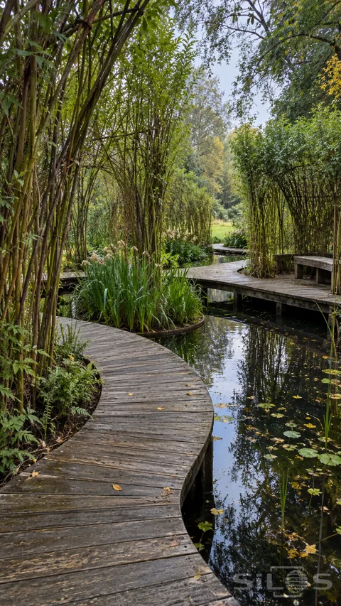Curving oak boardwalks and fitted benches wind between tall living willow screens and planted islands above a dark reflective pond.