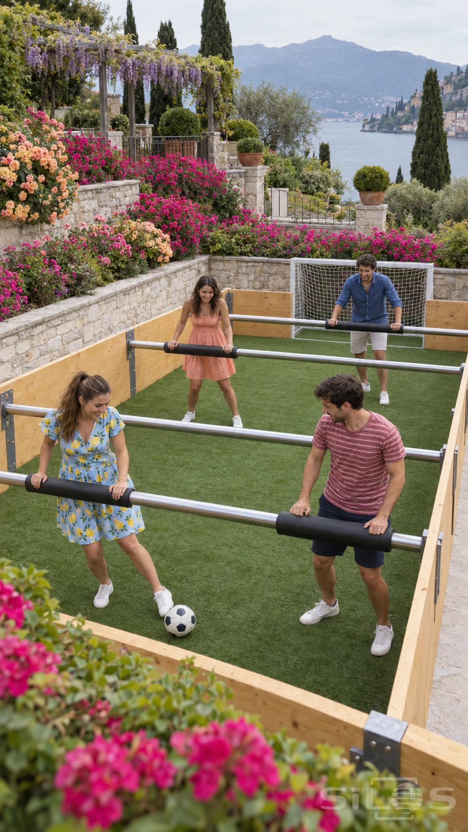 Four adults hold padded handles in a human foosball flower court while one woman taps a ball beneath planted terraces beside a lake.