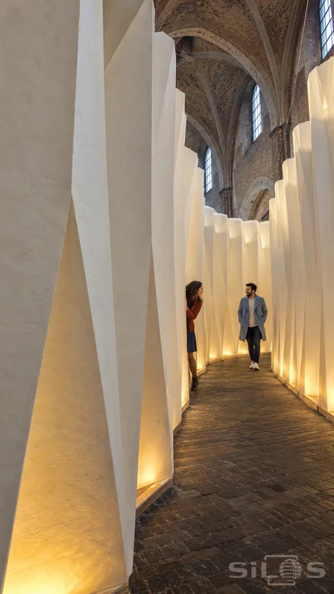 Two adults exchange a playful glance inside a paper canyon formed by tall illuminated pleated walls beneath a brick vault.