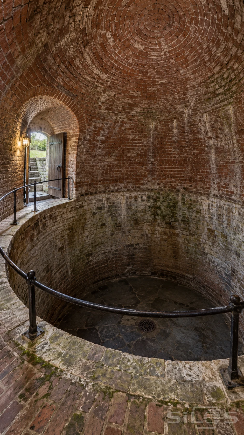 A brick icehouse vault encloses a circular chamber beneath an iron rail as daylight enters through an open oak door.