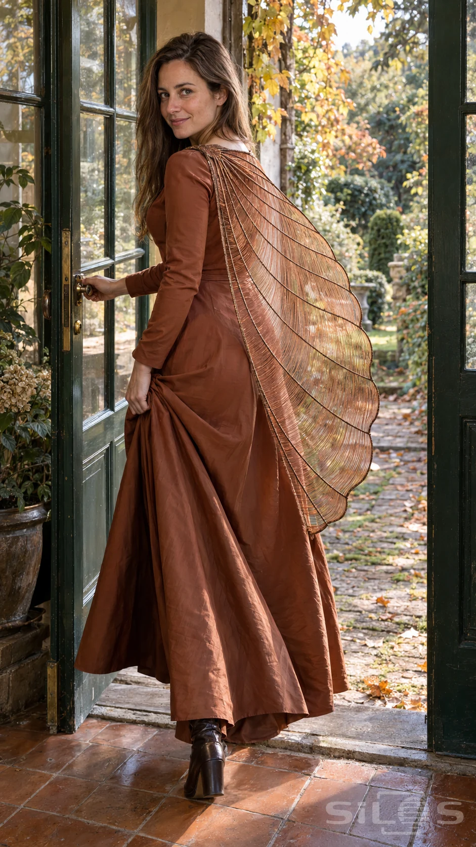 Woman in a russet silk dress turning through green garden doors with a sweeping transparent copper-thread fan cape.