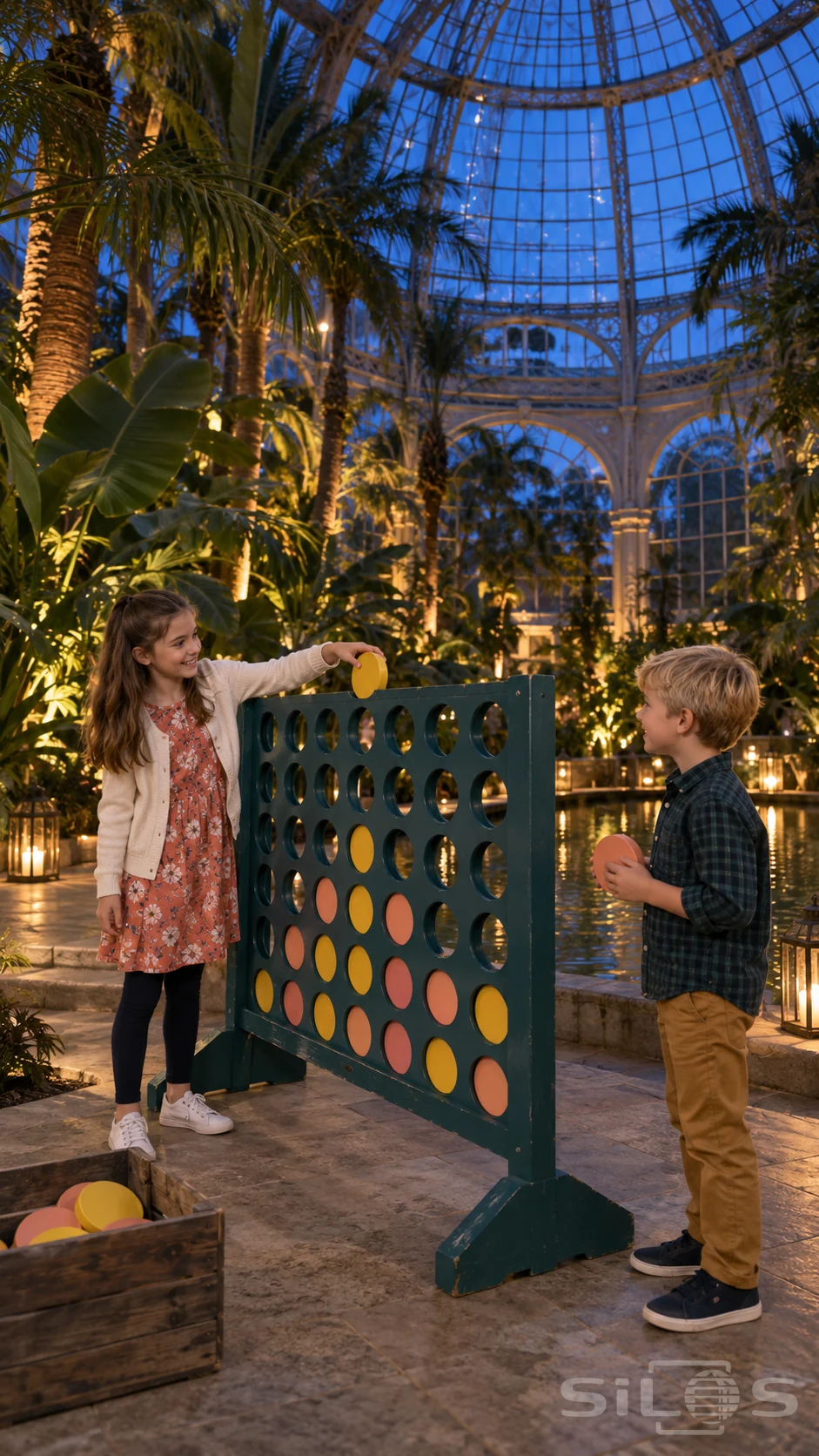 Two children play four in a row with yellow and coral wooden discs inside a tall palm dome illuminated at blue hour.