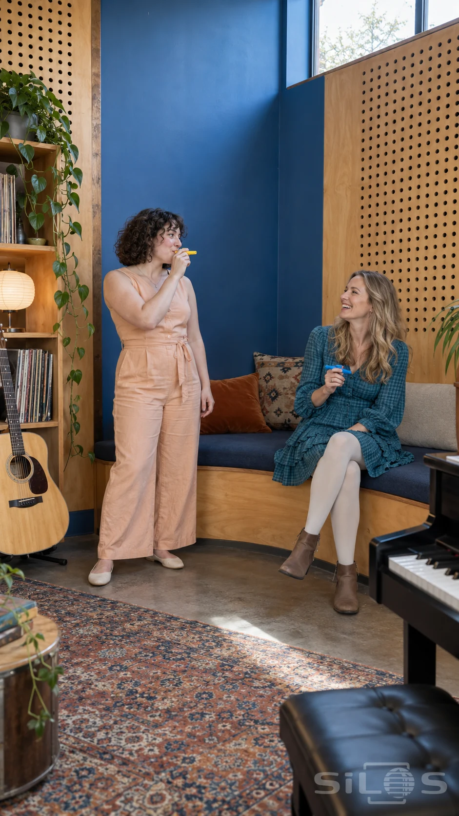 Two adult friends starting a kazoo duet in a blue alcove with perforated timber walls and a curved upholstered bench.