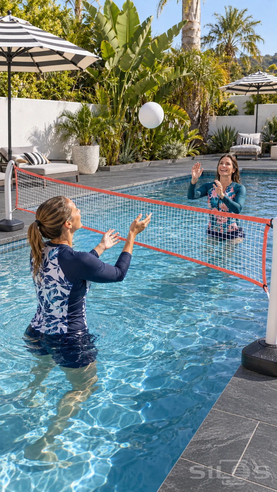 Two adult women play pool volleyball in leaf print swimwear beneath striped parasols and palms.