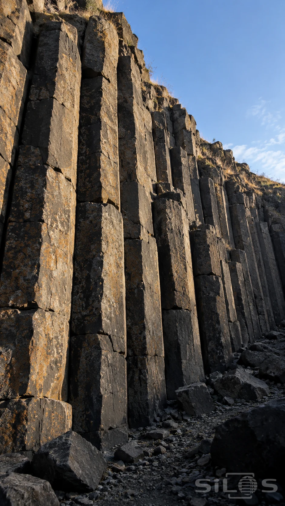 Tall basalt columns with horizontal fractures rise above broken stones, with dry grass along their tops and blue sky beyond.