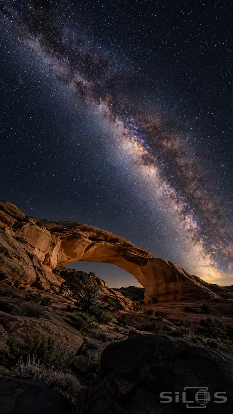 The Milky Way stretches diagonally above a low desert arch and dark foreground boulders.