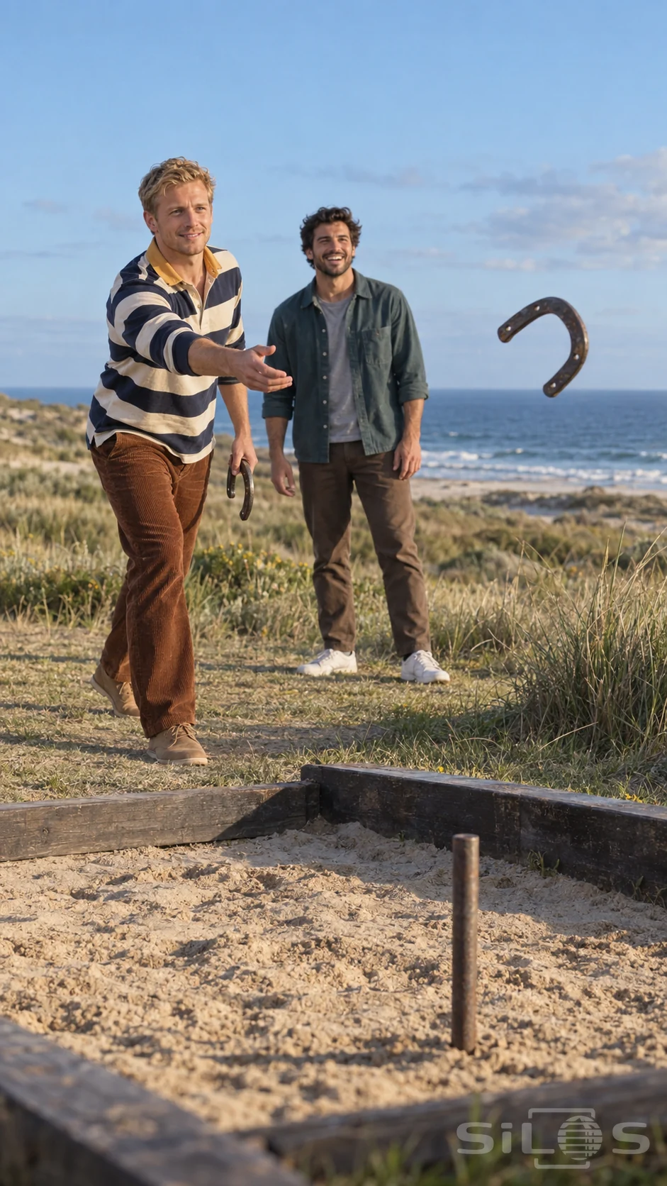 Two adult friends watch a horseshoe pitch toward a stake in a sandy pit beside the sea.
