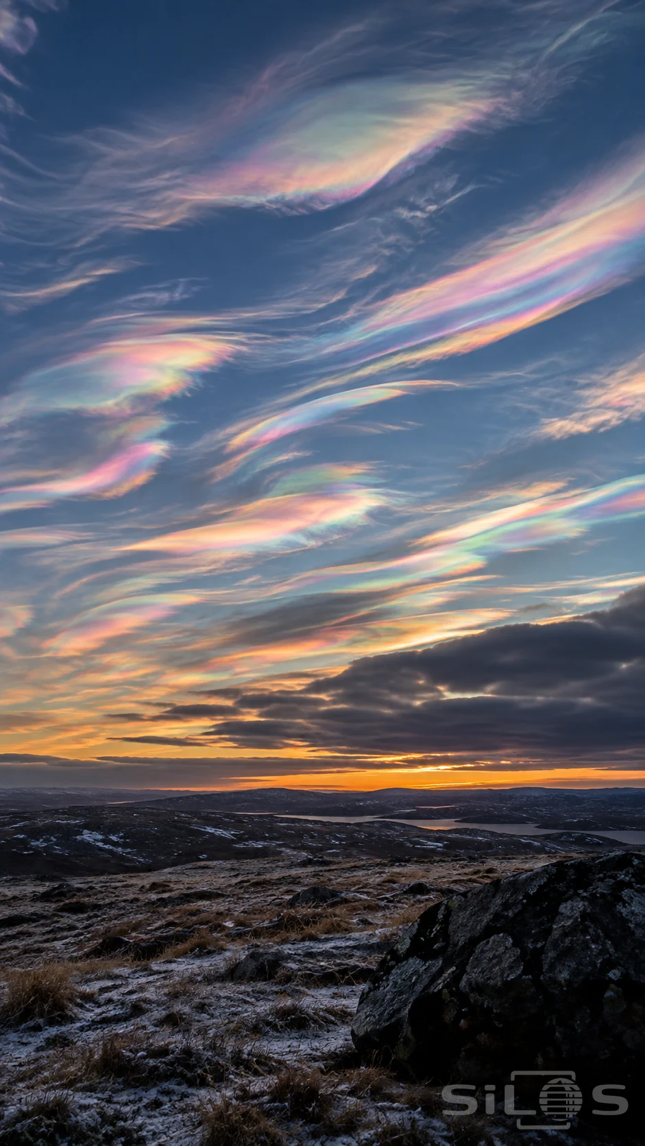 Nacreous clouds streak a slate-blue sky with pink and mint above a low frosty upland and dark foreground rock.
