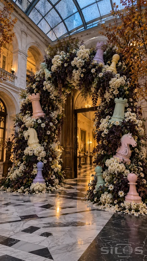 A tall arch of ivory flowers and dark foliage holds oversized pastel rooks; knights and pawns in a marble courtyard.