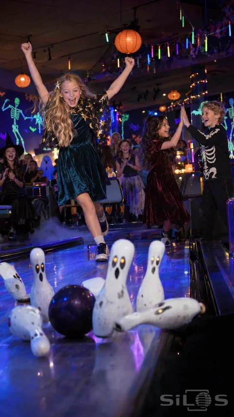 Children in teal and burgundy velvet dresses and a skeleton jumper celebrate beside blue bowling lanes; orange lanterns and ghost-painted pins.