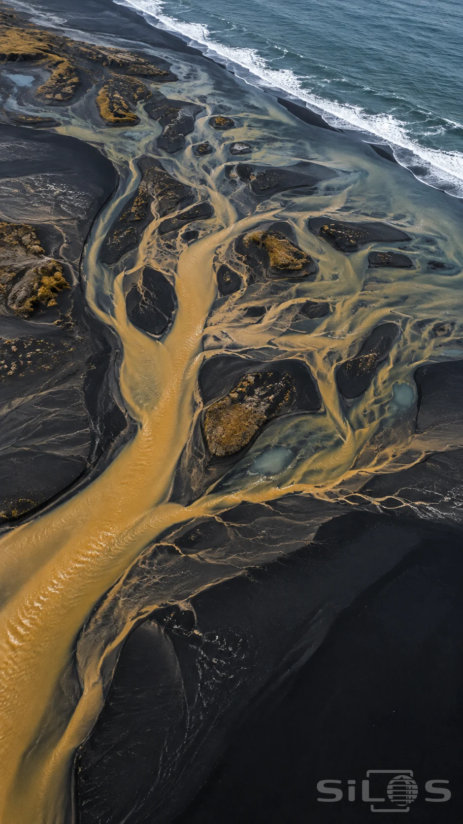An ochre braided river divides across blue-black sand beside dark islands and a diagonal strip of white ocean surf.