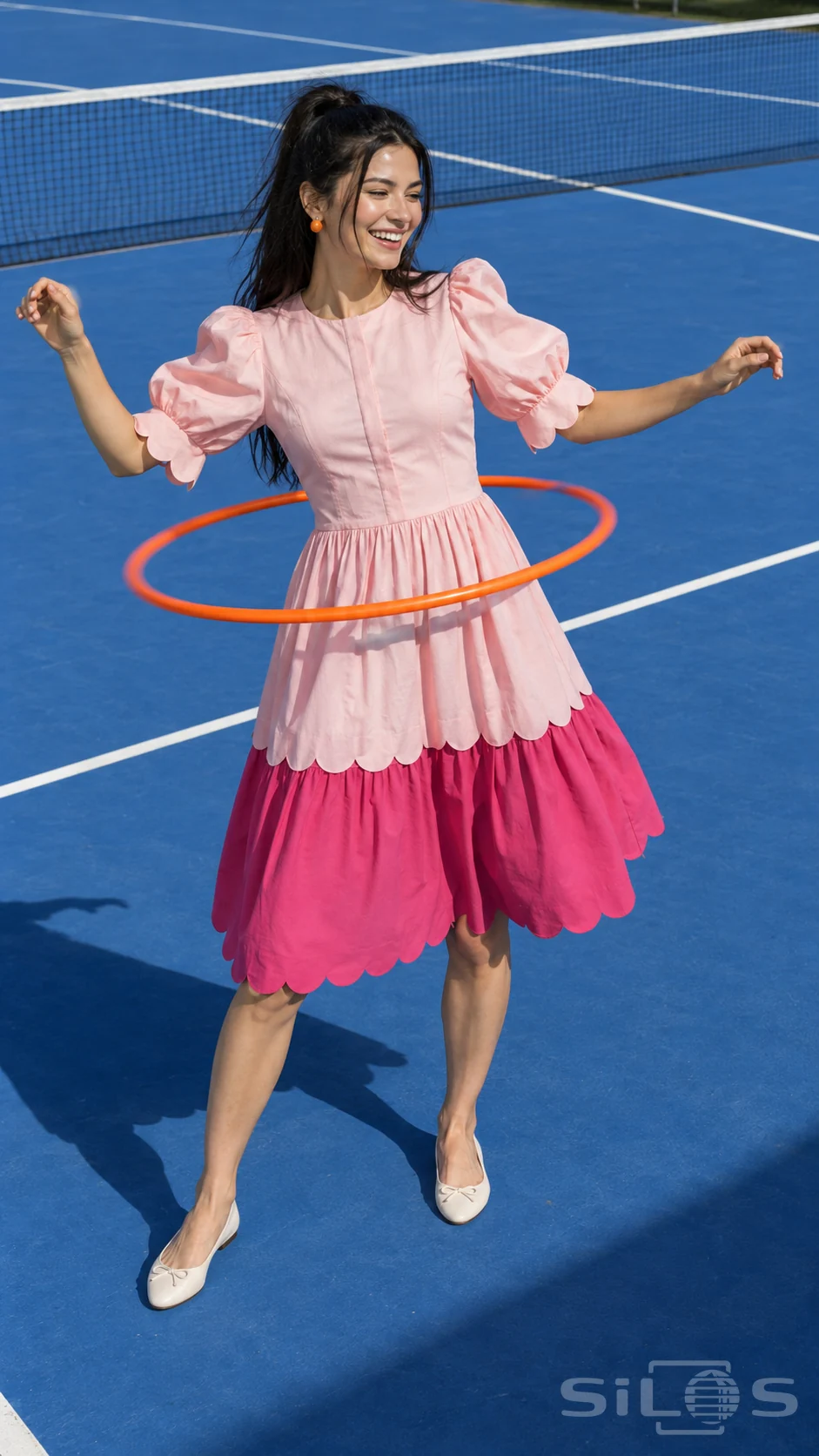 An adult woman in a pink tiered dress plays with an orange hula hoop on a blue tennis court in sunshine.
