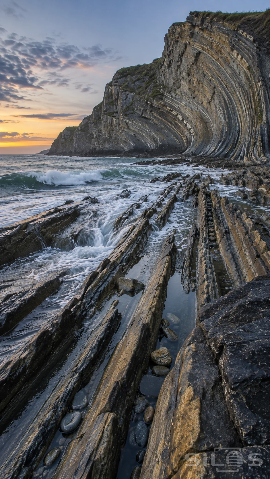Tidal flysch cliffs curve above long rock channels filled with pools and white surf at dusk.