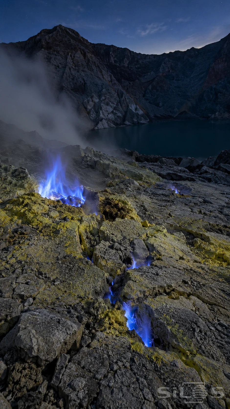 Blue sulfur flames rise from fissures in yellow mineral crust beside a dark crater lake.