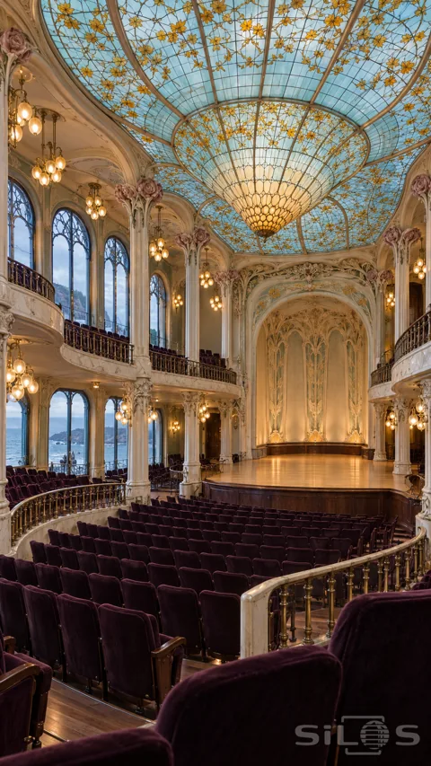 Empty concert hall with an aquamarine and amber stained-glass ceiling; cream floral columns; plum velvet seats and sea-facing windows.