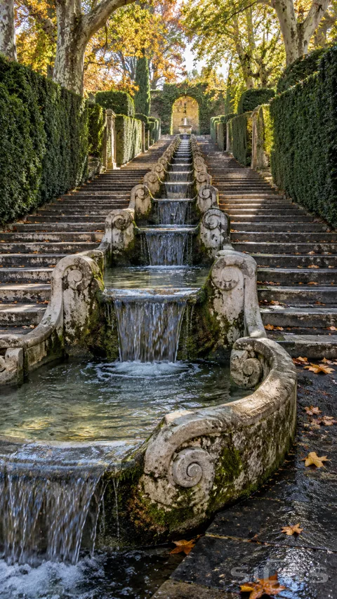 A long stone cascade with curled scroll edges descends between broad stairs and clipped hedges under golden autumn trees.