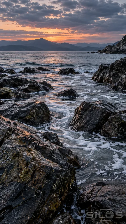 An empty rocky shore at dusk, with textured wet boulders and connected foam channels leading toward a quiet bay and layered mountain ridges.